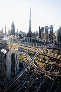 A futuristic cityscape featuring a network of highways intertwined with modern skyscrapers, with an iconic tall building dominating the skyline. The image showcases a blend of architectural designs, including glass facades and geometrically intricate structures. The scene is illuminated by soft daylight, enhancing the details of the urban landscape.