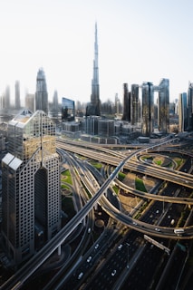 A futuristic cityscape featuring a network of highways intertwined with modern skyscrapers, with an iconic tall building dominating the skyline. The image showcases a blend of architectural designs, including glass facades and geometrically intricate structures. The scene is illuminated by soft daylight, enhancing the details of the urban landscape.