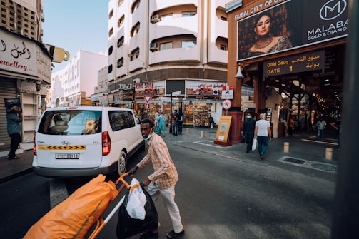 A bustling street scene featuring a pedestrian pushing a cart filled with bags beside a white van. The setting is a commercial area with various shops, including a spice shop. There's a prominent billboard advertising gold and diamond jewelry. The architecture is a mix of modern and traditional styles.