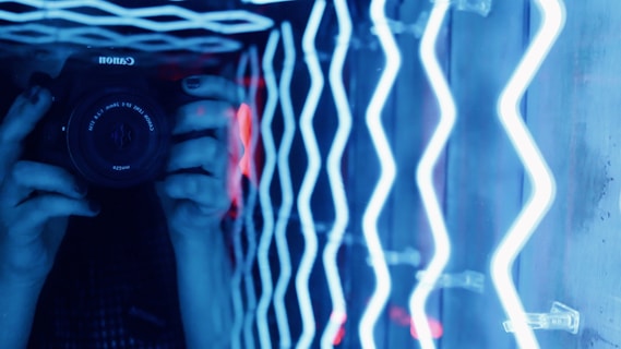A person holding a camera in front of a reflective surface adorned with bright, zigzagging neon blue lights. The person is only partially visible, with the focus on their hands gripping the camera. The lights create an abstract and vibrant backdrop, reflecting off the surface.