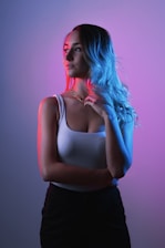 A person with long hair stands in a colorful lighting setup, with their hair flowing to one side. The lighting creates a dramatic effect with contrasting red and blue tones on either side of the person, enhancing the visual impact. They are wearing a white top and dark bottoms, and there is a contemplative expression on their face.