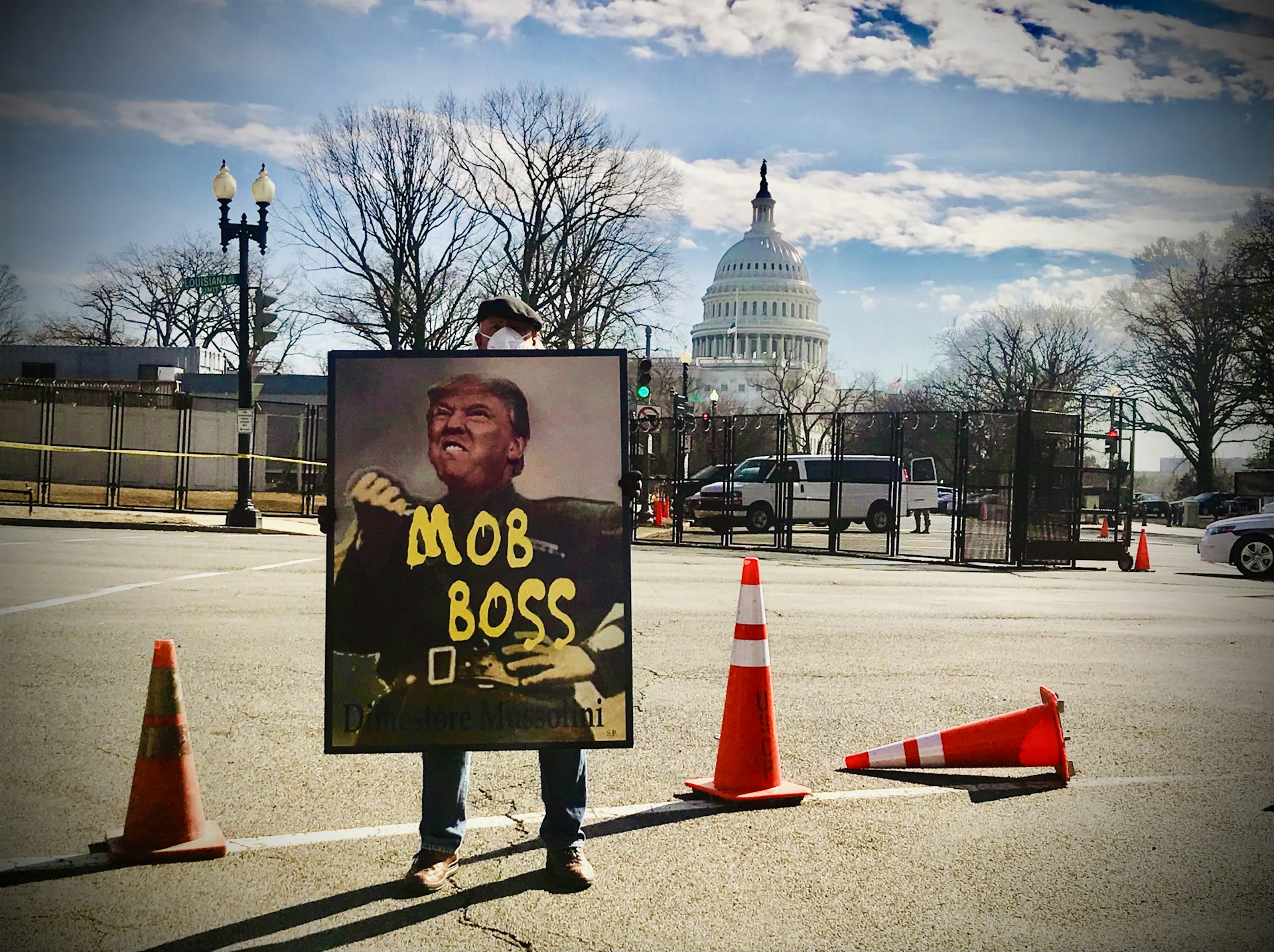 A person holds a large sign depicting a political figure with the words 'MOB BOSS' in front of the United States Capitol building. Orange traffic cones are set up around them on the street. The sky is partly cloudy, and trees without leaves are visible.