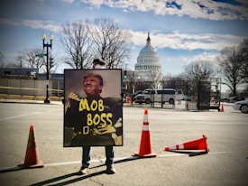 A person holds a large sign depicting a political figure with the words 'MOB BOSS' in front of the United States Capitol building. Orange traffic cones are set up around them on the street. The sky is partly cloudy, and trees without leaves are visible.