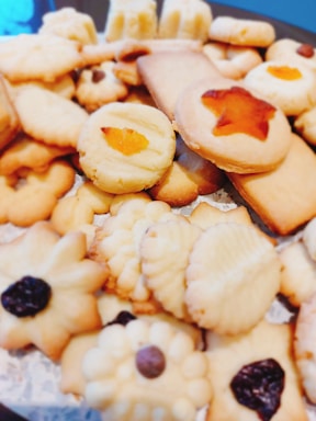 A variety of homemade cookies are arranged together. They include different shapes such as stars and flowers, with textures that appear soft and crumbly. Some cookies are topped with raisins or pieces of fruit, adding visual appeal.