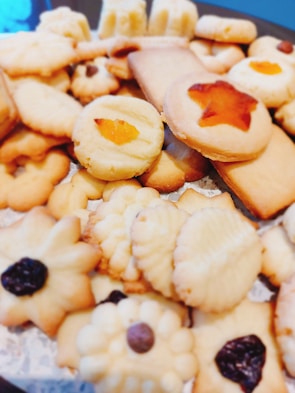 A variety of homemade cookies are arranged together. They include different shapes such as stars and flowers, with textures that appear soft and crumbly. Some cookies are topped with raisins or pieces of fruit, adding visual appeal.