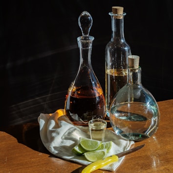 Three glass bottles with cork stoppers are displayed on a wooden table, accompanied by a small glass filled with liquid. A white cloth is spread out with lime wedges and a knife resting nearby. The scene is softly lit, casting subtle reflections and shadows on the surface.