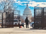 A security checkpoint with metal fencing and a guard accompanied by a dog in the foreground. The U.S. Capitol building is prominently visible in the background under a bright blue sky with scattered clouds. Bare trees and various vehicles are also part of the scene.