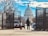A security checkpoint with metal fencing and a guard accompanied by a dog in the foreground. The U.S. Capitol building is prominently visible in the background under a bright blue sky with scattered clouds. Bare trees and various vehicles are also part of the scene.