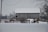 A snow-covered rural setting features a large wooden barn with a metal roof. A red wooden fence runs across the foreground with a small gate. Just outside the barn, farm equipment, including a satellite dish and some machinery, is partially covered in snow. Leafless trees and a muted, overcast sky complete the wintery scene.