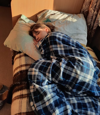 A person is sleeping comfortably on a bed, wrapped in a blue and white plaid blanket. The room is warmly lit by sunlight streaming in, creating a cozy and peaceful atmosphere. A decorative pillow is placed at the head of the bed, and there is also a patterned curtain visible in the background.