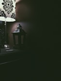 A dimly lit room featuring a decorative table lamp with a black and white pattern on the lampshade. Next to the lamp is a small, dark lantern sitting on a wooden surface. The right side of the image is mostly in shadow, creating a contrast with the light illuminating the left side.