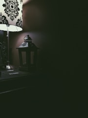 A dimly lit room featuring a decorative table lamp with a black and white pattern on the lampshade. Next to the lamp is a small, dark lantern sitting on a wooden surface. The right side of the image is mostly in shadow, creating a contrast with the light illuminating the left side.