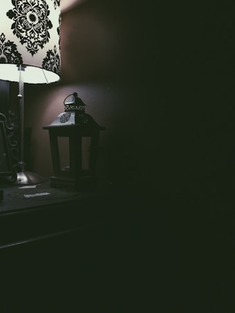 A dimly lit room featuring a decorative table lamp with a black and white pattern on the lampshade. Next to the lamp is a small, dark lantern sitting on a wooden surface. The right side of the image is mostly in shadow, creating a contrast with the light illuminating the left side.