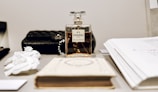 A bottle of Chanel No. 5 perfume is positioned on a table among other elegant items. To the left, there are two white decorative flowers. In the background, a black quilted leather bag is visible. On the right side, there are some open books with handwritten notes visible, lending a sophisticated ambiance to the scene.