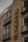 A building facade featuring a prominent vertical sign for a hotel. The sign displays the word 'HOTEL' in bold, yellow letters against a dark background. Below the word, there is a smaller circular section with the words 'Suites Real'. The building has multiple windows with decorative wrought iron railings on the balconies.
