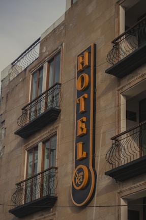 A building facade featuring a prominent vertical sign for a hotel. The sign displays the word 'HOTEL' in bold, yellow letters against a dark background. Below the word, there is a smaller circular section with the words 'Suites Real'. The building has multiple windows with decorative wrought iron railings on the balconies.