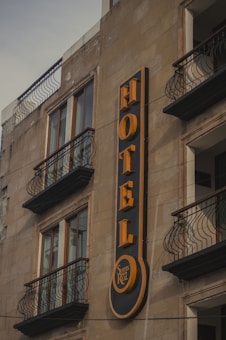 A building facade featuring a prominent vertical sign for a hotel. The sign displays the word 'HOTEL' in bold, yellow letters against a dark background. Below the word, there is a smaller circular section with the words 'Suites Real'. The building has multiple windows with decorative wrought iron railings on the balconies.