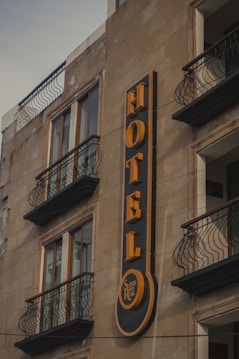 A building facade featuring a prominent vertical sign for a hotel. The sign displays the word 'HOTEL' in bold, yellow letters against a dark background. Below the word, there is a smaller circular section with the words 'Suites Real'. The building has multiple windows with decorative wrought iron railings on the balconies.