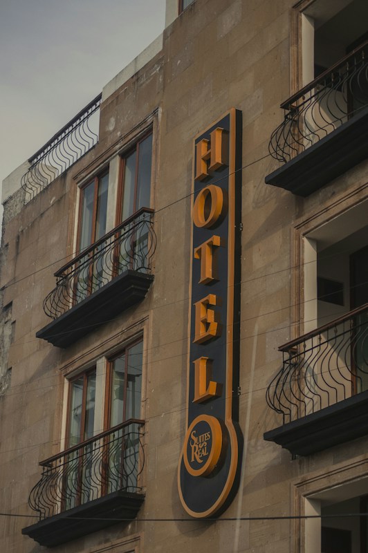 A building facade featuring a prominent vertical sign for a hotel. The sign displays the word 'HOTEL' in bold, yellow letters against a dark background. Below the word, there is a smaller circular section with the words 'Suites Real'. The building has multiple windows with decorative wrought iron railings on the balconies.