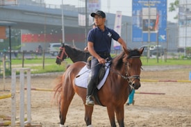 A person wearing a blue polo shirt and black cap is sitting on a brown horse with a white saddle. They are holding the reins and appear to be in an outdoor practice area with another horse and rider in the background. The area is fenced and there are several infrastructure elements and buildings visible in the distance.