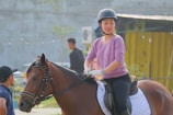 A person wearing a purple sweater and helmet is riding a brown horse. Another person nearby is wearing a cap and appears to be giving instructions or having a conversation. The setting includes a concrete wall and outdoor area with string lights and plants.