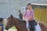 A person wearing a purple sweater and helmet is riding a brown horse. Another person nearby is wearing a cap and appears to be giving instructions or having a conversation. The setting includes a concrete wall and outdoor area with string lights and plants.