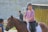 A person wearing a purple sweater and helmet is riding a brown horse. Another person nearby is wearing a cap and appears to be giving instructions or having a conversation. The setting includes a concrete wall and outdoor area with string lights and plants.