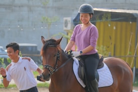 A person wearing a helmet and purple sweater is enjoying a horseback ride, smiling and looking relaxed. Another individual in a white polo shirt is walking beside the horse, also appearing cheerful. The background includes a yellow structure and greenery.