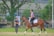 A person wearing a helmet and boots is riding a brown horse with protective gear. Another person stands nearby with folded arms, observing. The setting is an outdoor area with green grass, trees, and a fence, possibly a training ground for horses.