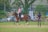 A person is riding a brown horse in an open grassy area, wearing equestrian gear including a helmet. Another individual stands nearby, observing with hands on hips. In the background, there are parked cars and motorcycles, along with lush green trees and a building.