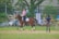 A person is riding a brown horse in an open grassy area, wearing equestrian gear including a helmet. Another individual stands nearby, observing with hands on hips. In the background, there are parked cars and motorcycles, along with lush green trees and a building.