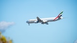 A large commercial airplane with the Emirates logo is flying against a clear blue sky. The aircraft features distinct markings on the tail and appears to be preparing for landing as the landing gear is extended.