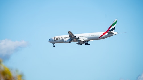 A large commercial airplane with the Emirates logo is flying against a clear blue sky. The aircraft features distinct markings on the tail and appears to be preparing for landing as the landing gear is extended.