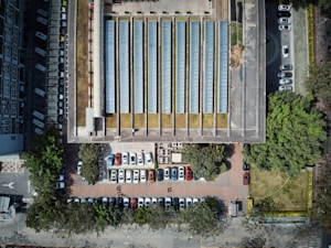 An aerial view of a parking lot adjacent to a large building with a series of long, narrow glass-covered structures resembling skylights or solar panels. Multiple cars are parked in organized rows, surrounded by green trees and well-maintained grass areas.