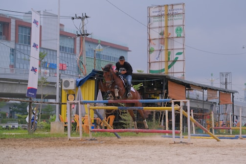 A man is riding a horse as it jumps over a barrier in an outdoor equestrian arena. Behind them are several buildings, one with a large advertisement and another with modern architecture. The ground is sandy and there are several colorful barriers around.