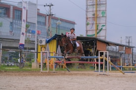 A person is riding a horse, jumping over an obstacle in an outdoor equestrian setting. The background features an urban environment with buildings, including a large advertisement and signs. Various equipment and objects are visible around the jumping area, such as chairs, a tent, and barriers.
