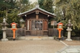 A traditional wooden shrine stands amid a tranquil setting, surrounded by lush greenery and fenced with a wooden picket fence. The architecture features a peaked roof, and there are two stone lanterns flanking the entrance. Bright orange-lacquered lanterns, enhanced with decorative elements, add vibrant contrast.