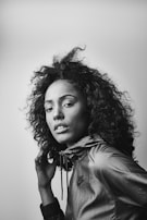 A black and white portrait of a person with curly hair wearing a sporty jacket. The subject is looking confidently at the camera with a hand near their face.