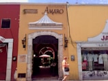 A quaint street scene featuring a yellow building with a restaurant entrance labeled 'AMARO'. The facade includes an ornate white canopy and two traditional lanterns flanking the doorway. To the left, a red building hosts other businesses, and to the right, mannequins displayed in a shop window selling clothes. A person is walking past the entrance, dressed casually and holding items in their hands.