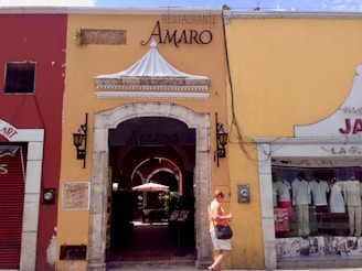 A quaint street scene featuring a yellow building with a restaurant entrance labeled 'AMARO'. The facade includes an ornate white canopy and two traditional lanterns flanking the doorway. To the left, a red building hosts other businesses, and to the right, mannequins displayed in a shop window selling clothes. A person is walking past the entrance, dressed casually and holding items in their hands.