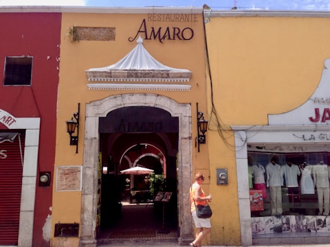 A quaint street scene featuring a yellow building with a restaurant entrance labeled 'AMARO'. The facade includes an ornate white canopy and two traditional lanterns flanking the doorway. To the left, a red building hosts other businesses, and to the right, mannequins displayed in a shop window selling clothes. A person is walking past the entrance, dressed casually and holding items in their hands.