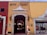 A quaint street scene featuring a yellow building with a restaurant entrance labeled 'AMARO'. The facade includes an ornate white canopy and two traditional lanterns flanking the doorway. To the left, a red building hosts other businesses, and to the right, mannequins displayed in a shop window selling clothes. A person is walking past the entrance, dressed casually and holding items in their hands.