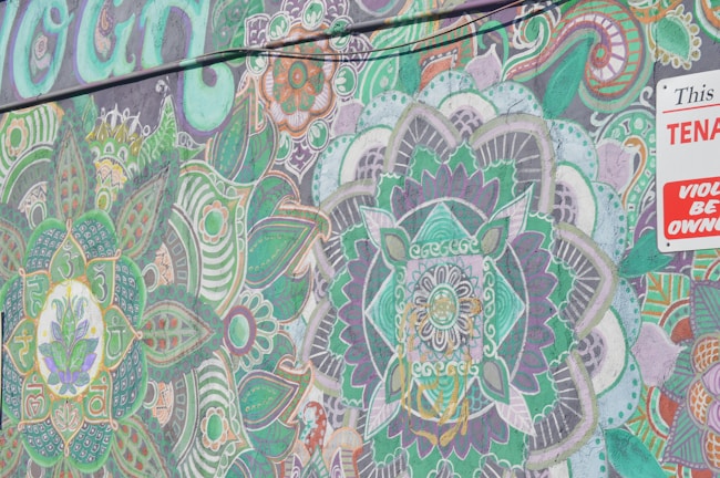 The image features a vibrant mural with intricate mandala patterns and swirling floral designs. Colors range from various shades of green to hints of purple, orange, and white. There is a bold, partially visible sign in the top right corner with contrasting red and white colors.