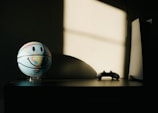 A basketball featuring a world map and a smiley face stands on a table, illuminated by a soft light from a window. Next to it, there is a game controller and a modern gaming console with a sleek design. The background has a shadow cast on the wall, adding depth and contrast to the scene.