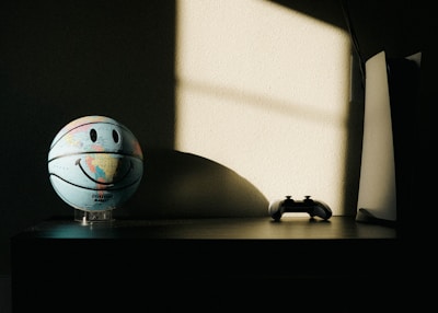 A basketball featuring a world map and a smiley face stands on a table, illuminated by a soft light from a window. Next to it, there is a game controller and a modern gaming console with a sleek design. The background has a shadow cast on the wall, adding depth and contrast to the scene.