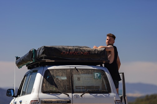 A man is standing next to a white Toyota Land Cruiser with a large black cargo bag secured on its roof. The vehicle is parked outdoors with a clear blue sky and distant mountains in the background. The man is wearing a black sleeveless shirt.