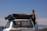 A man is standing next to a white Toyota Land Cruiser with a large black cargo bag secured on its roof. The vehicle is parked outdoors with a clear blue sky and distant mountains in the background. The man is wearing a black sleeveless shirt.