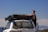 A man is standing next to a white Toyota Land Cruiser with a large black cargo bag secured on its roof. The vehicle is parked outdoors with a clear blue sky and distant mountains in the background. The man is wearing a black sleeveless shirt.