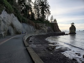 A coastal walking path runs alongside a rocky cliff covered in greenery, with tall trees silhouetted against the sky. The path curves around the coastline, leading towards a solitary rock formation standing in the water. Several people are seen walking or cycling along the path, enjoying the scenic view.