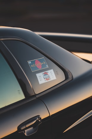 The image captures a close-up view of a car, focusing on the window area. There are stickers visible on the window, which include some text and a graphic of a vehicle. The car is black and the photo is taken at sunset, casting a warm, golden glow on the vehicle's surface.